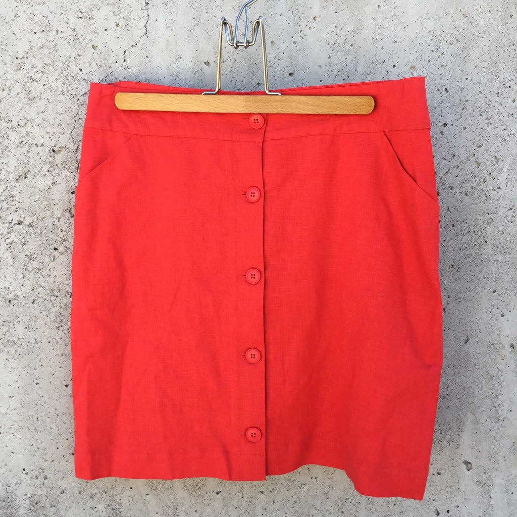 Jupe rouge longueur aux genoux, boutonnée de haut en bas à l’avant, doublure rouge, lien dans le dos, 2 poches à l’avant.