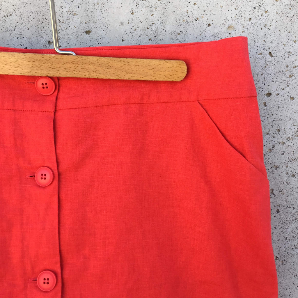 Jupe rouge longueur aux genoux, boutonnée de haut en bas à l’avant, doublure rouge, lien dans le dos, 2 poches à l’avant.