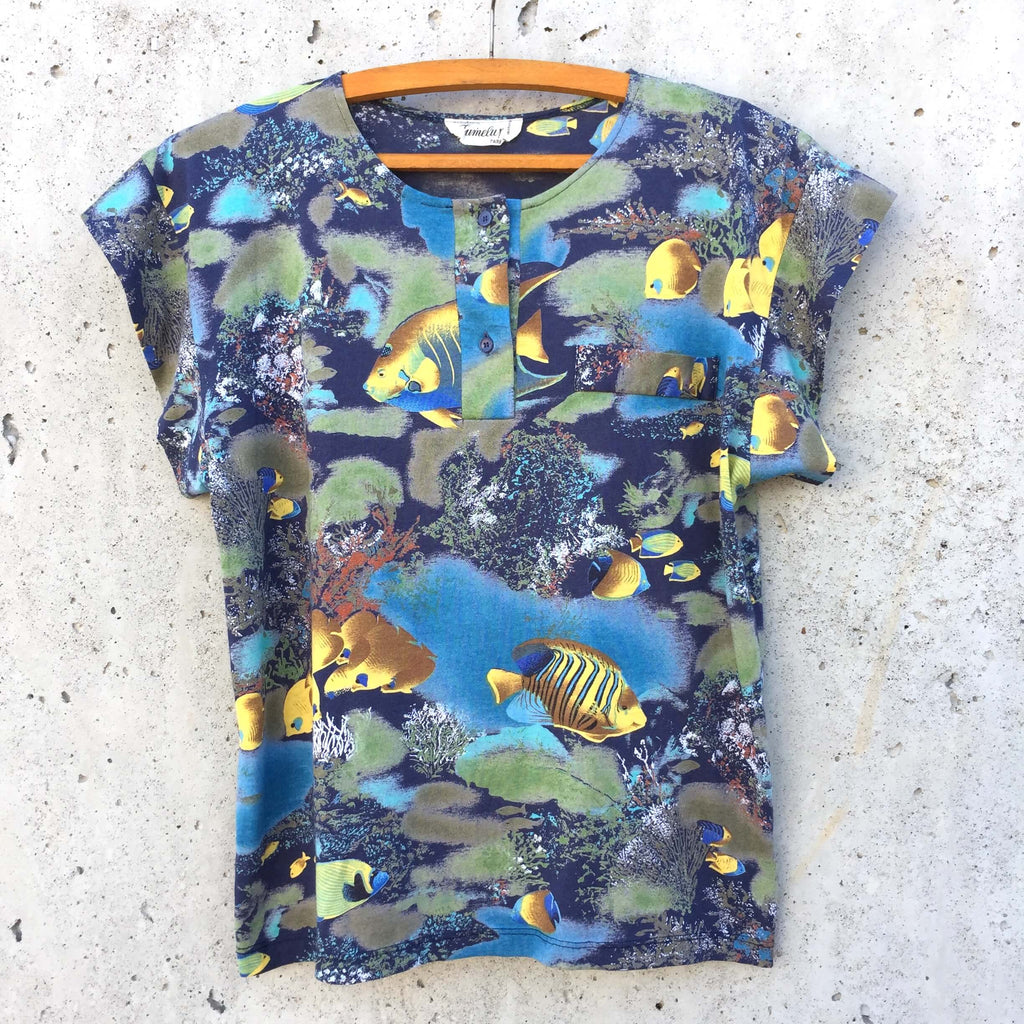 Tee-shirt à motif aquarium, dominante de couleurs bleues et vertes, et poissons jaunes. Col boutonné, fausse poche sur le devant.
