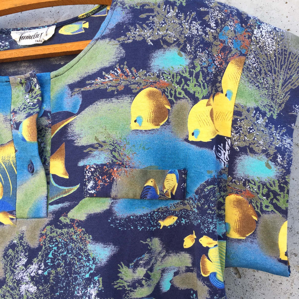 Tee-shirt à motif aquarium, dominante de couleurs bleues et vertes, et poissons jaunes. Col boutonné, fausse poche sur le devant.